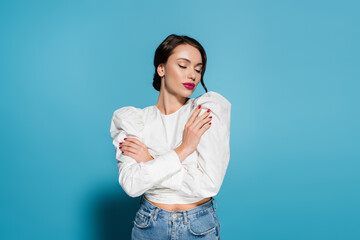 charming young woman in white blouse posing with crossed arms and closed eyes isolated on blue.