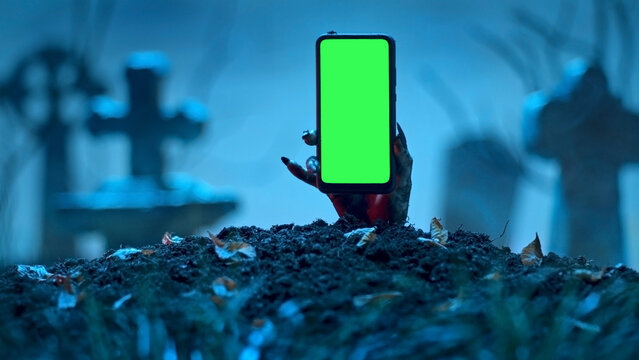 Zombie Hand Rising Up Smartphone With Green Screen Out Of Grave. Holiday Event Halloween Concept.