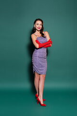 full length of brunette young woman in red gloves and purple dress smiling while posing with crossed arms on green.