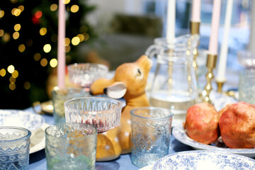 The festive dinner table stands against the backdrop of a Christmas tree. On the table there is a blue tablecloth, plates, glasses and decor