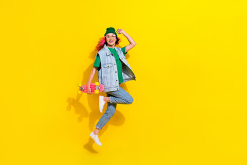 Full length photo of sweet positive lady wear jeans waistcoat spectacles jumping high holding skateboard isolated yellow color background
