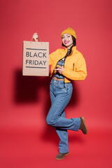 full length of happy young woman in beanie hat and puffer jacket pointing at shopping bag with...