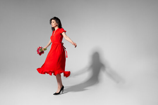 Full lengtu view of a beautiful brunette young woman with makeup in red dress holding a bouquet of tulips and spins, over grey background.