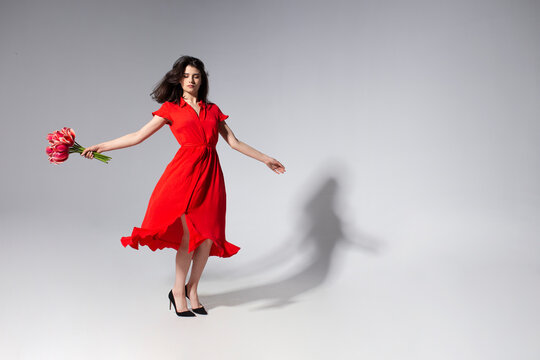 Full lengtu view of a beautiful brunette young woman in red dress holding a bouquet of tulips and spins, over grey background.