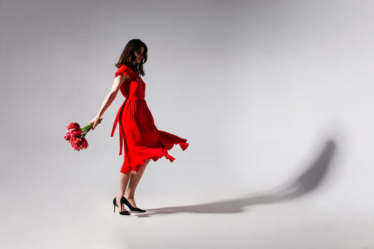 Full lengt view of a sensual brunette young woman in red dress holding a bouquet of tulips and spins, isolated grey background. Copy space.
