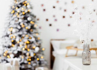 A vase with decorative plant branches stands on a table in the bedroom against the backdrop of Christmas lights.