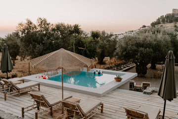 The relaxing scene of a swimming pool area, sun umbrella, trees and loungers at sunset in Rhodes Island, Greece