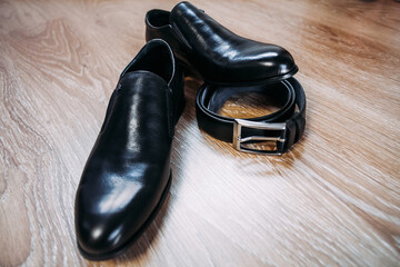 Black men's patent leather shoes for the groom and a leather black belt with an iron buckle lie on a wooden brown textured surface.