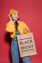 happy young woman in orange beanie hat and puffer jacket holding shopping bag with black friday...