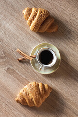 A cup of hot fragrant black coffee in a saucer with croissants and cinnamon sticks, on a light brown wooden table.