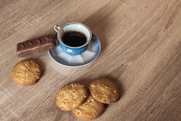 A cup of hot fragrant black coffee in a saucer with cookies and a chocolate bar, on a wooden light brown textured table.