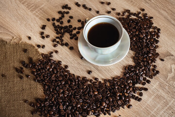 A cup of hot aromatic coffee in a saucer. Coffee beans are scattered on a light brown wooden table.