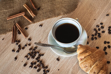 A cup of hot coffee in a plate with a spoon and a croissant. Coffee beans and cinnamon are scattered on a wooden light brown table.