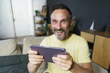 Excited playing games online middle aged man use digital tablet sitting on the sofa at home wearing casual. Mature freelancer man hold digital tablet work from home. Business online