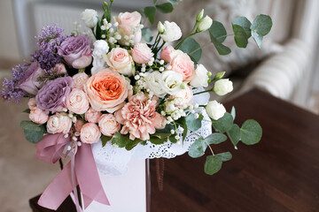 A chic festive bouquet of flowers, made of pastel-colored roses, in a decorative paper box decorated with pink ribbons, stands on a table in a bright hotel room, during the day.