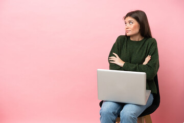 Young caucasian woman sitting on a chair with her laptop isolated on pink background making doubts gesture while lifting the shoulders