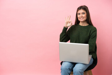 Young caucasian woman sitting on a chair with her laptop isolated on pink background showing ok sign with fingers