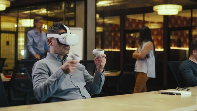 Male office worker wearing VR headset and using wireless controllers, gesturing, watching data in virtual reality. Work in modern office of hi-tech company. Cyberspace digital technology