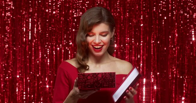 Surprised woman opening christmas gift box with magic shining light on red shiny backdrop foil curtain background