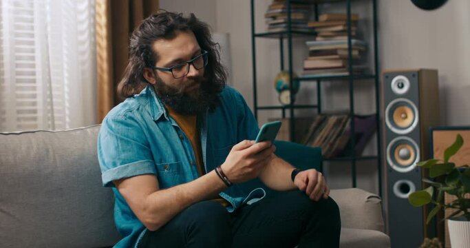 A bearded handsome guy is sitting on the couch with his dog in the living room. The bearded man is browsing insurance policy quotes on smartphone.