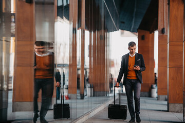 Fototapeta premium Going to airport terminal. Confident businessman traveler walking on city streets and pulling his suitcase drinking coffee and speaking on smartphone