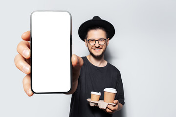 Studio portrait of young happy man holding cardboard holder with papers cups of coffee, showing big smartphone with mockup, empty blank on screen, on white background.