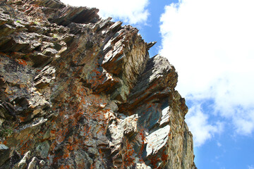 Fragment of a large gray-yellow rock with peaked ledges and red lichen against a sky with clouds, bottom view, summer, sunny