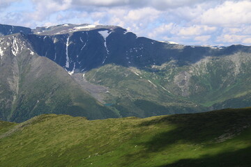 The slopes of huge mountains with grass and rocks in summer in sunny weather, on the far slope you can see a lake, in some places there is snow, a sky with clouds