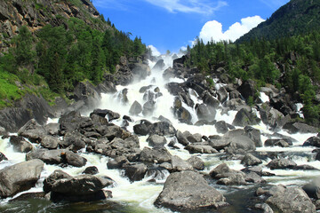A powerful stream of water from the Uchar waterfall rushes over huge stones from the mountain, coniferous trees grow on the rocky slopes of the mountains, a clear sunny day, the sky with clouds