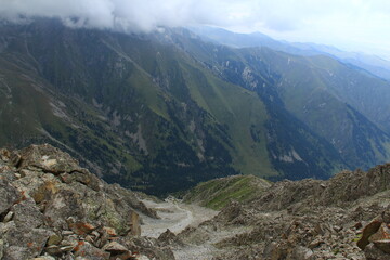 A mountain trail descends down into the valley, stones and rocks surround the trail, trees and grass grow on the side of the mountain, the sky is covered with clouds, a summer cloudy day is high in th