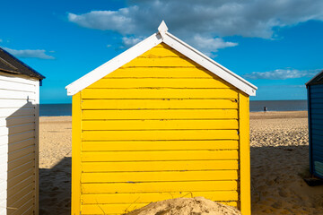 Naklejka premium Yellow painted wooden beach hut seen on a sandy beach on a famous beach area in Suffolk, UK. A couple can be seen walking along the shore in the distance.