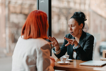 Two young business women sitting at table in cafe. Girl shows colleague information on laptop screen. Girl using smartphone, blogging. Teamwork, business meeting.