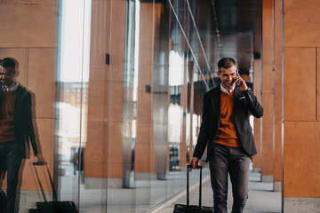 Going to airport terminal. Confident businessman traveler walking on city streets and pulling his suitcase drinking coffee and speaking on smartphone