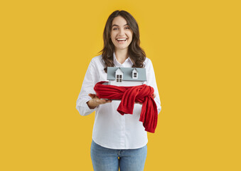 Joyful woman holds toy house wrapped in warm scarf as symbol for heating system or cold snowy...