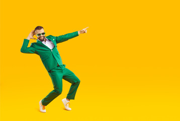 Fototapeta na wymiar Full body happy funny smiling joyful handsome young man in trendy stylish green suit and sunglasses having fun, dancing isolated on yellow color background and pointing finger away at blank copy space
