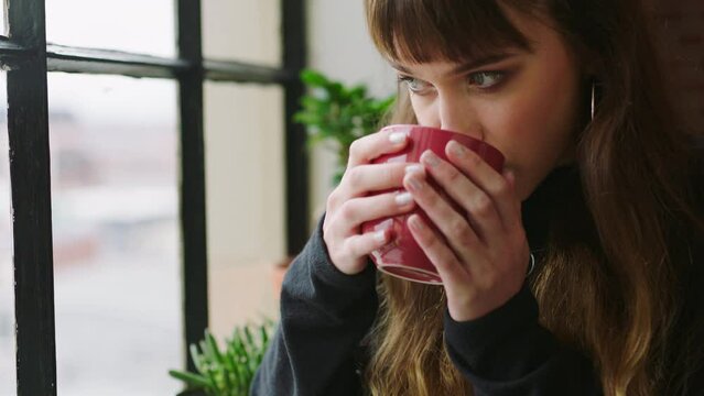 Thinking, window and sad woman drink tea while looking nostalgic female standing by the windowsill holding a cup of coffee and remember memory. Lady having a warm cozy beverage think of grief or fail