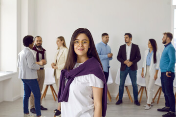 Fototapeta premium Portrait of young corporate business leader or intern girl. Happy good looking long haired brunette woman in eyeglasses and casual outfit standing in office with team of employees in background