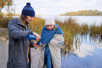 Holidays with friend. Young couple drink hot beverage from thermos near river.