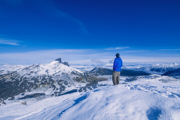 Adventurous athletic male hiker standing in snow at the summit of Panorama Ridge looking at The Black Tusk on a beautiful sunny day.
