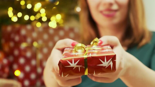 Red-haired woman gives present decorated with golden ribbon to friend near Christmas tree. Ginger woman wearing green shirt excited to celebrate Christmas