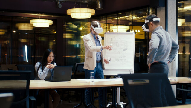 Multi-ethnic engineers in VR headsets, using laptop and wireless controllers, examining blueprint, watching data, working in modern office of hi-tech company. Cyberspace digital technology