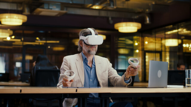Male office worker wearing VR headset and using wireless controllers and laptop, gesturing, watching data in virtual reality. Work in modern office of hi-tech company. Cyberspace digital technology - Powered by Adobe