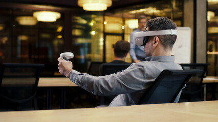 Back view of man wearing VR headset and using wireless controllers, watching data in virtual reality. Work in modern office of hi-tech company with diverse coworkers. Cyberspace digital technology