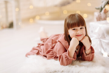 Fototapeta premium little cute child girl in pink dress lying on white furry carpet in decorated Christmas room interior and looking in camera. Beautiful bokeh. Happy family New Year, Xmas concept