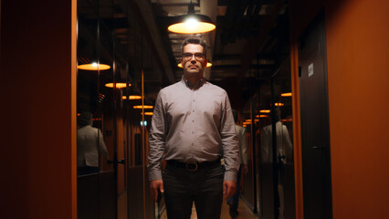 Fototapeta na wymiar Happy male office worker in suit and glasses standing in office corridor, smiling, looking at camera. Successful mature businessman in hallway of marketing company modern business center