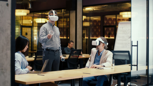 Multi-ethnic office workers testing VR headsets together, using tablet computer and wireless controllers, watching data, working in modern office of hi-tech company. Cyberspace digital technology
