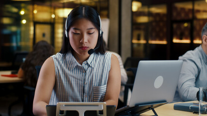 Asian woman in headset talking with client, using laptop and digital tablet. Female operator working in call center office of marketing company