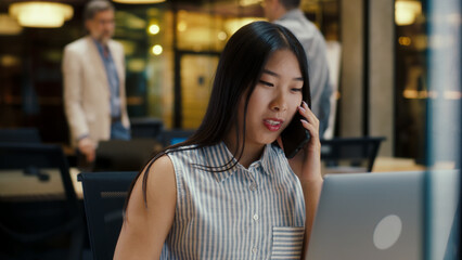 Asian woman talking with client by phone while working in modern office near colleagues, using laptop and digital tablet. Business woman discussing e-commerce startup. Strategy of the company