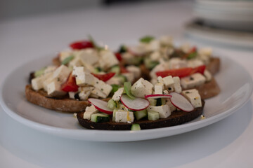 italian bread with greek cheese, tomatoes, cucumbers, radishes and basil and with various spices, on a white plate and a white kitchen table.