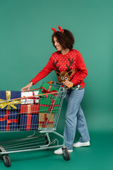 african american woman in deer horns headband taking present from shopping cart on green background